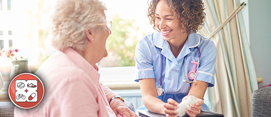 Young nurse holding a pill bottle and talking to older lady in a home. Both are seated.