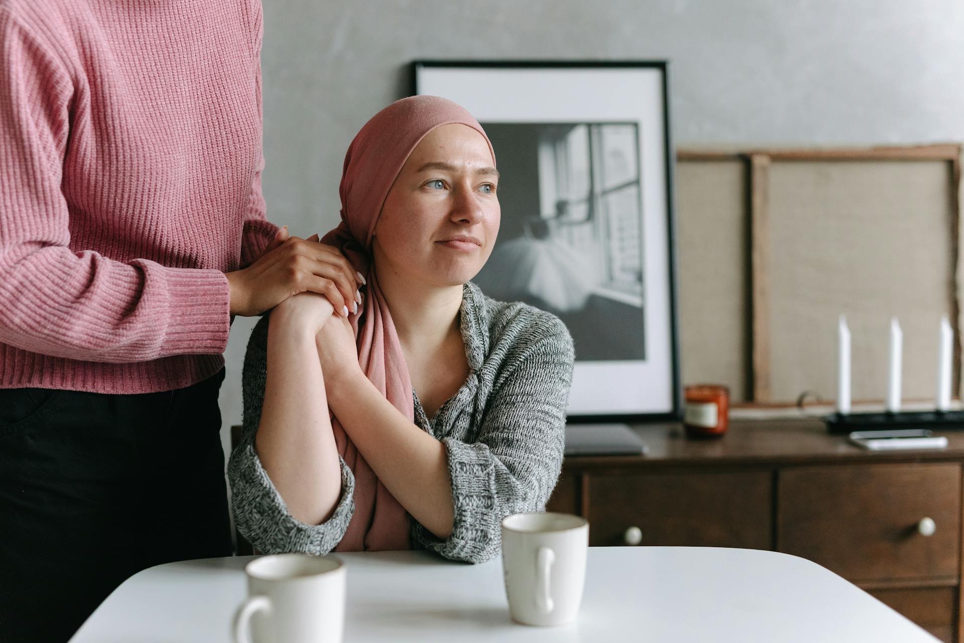 Seated woman with headscarf holding hand of person standing beside her.